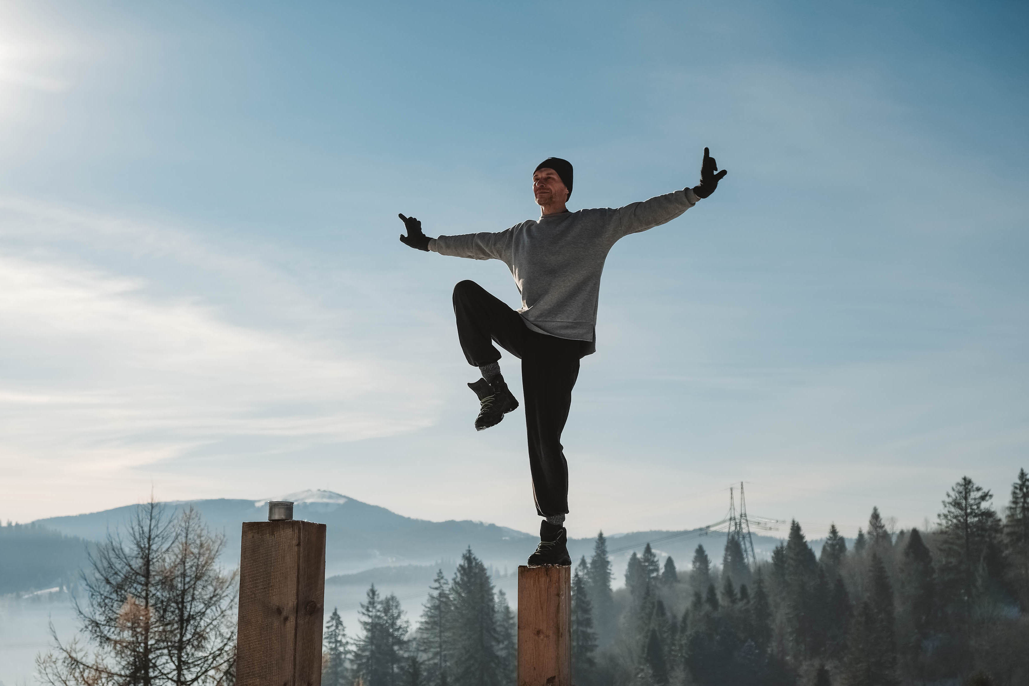 Athletic man doing a pose at the top of a wooden pole, arms extended, wearing a beanie